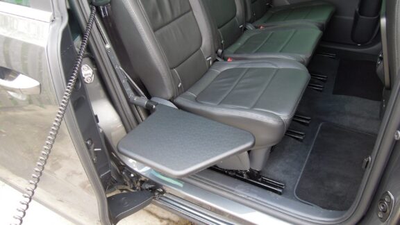 An electric transfer plate sits at seat level next to a row of seats in a minivan next to the sliding door. An corded wire is attached to the mechanism that raises and lowers the plate.