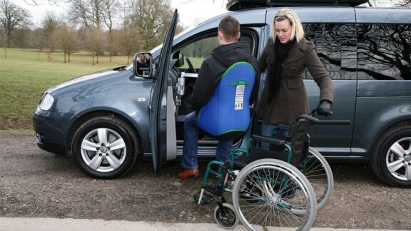 A person is lifted using a blue seat harness from their wheelchair and into the passenger side of a VW van.