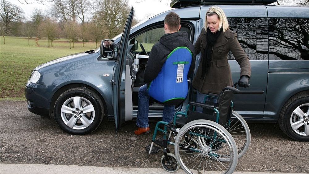 A person is lifted using a blue seat harness from their wheelchair and into the passenger side of a VW van.