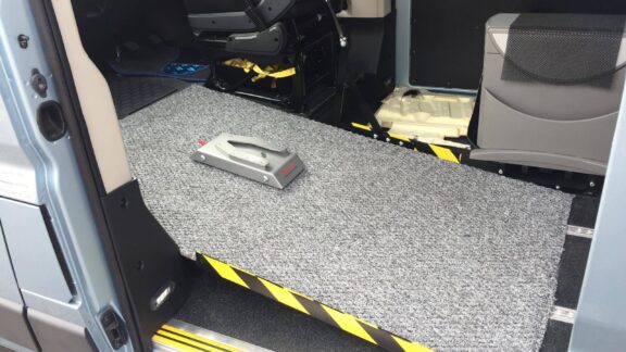 Wheelchair lockdown shown in a van, in the centre of a carpeted panel is a metal slot which the wheelchair pin affixes to.