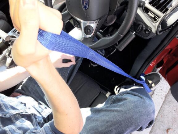 The Leg Lifter is shown in use, a blue strap from the driver's foot is being pulled with their hand to help move their leg into the car.