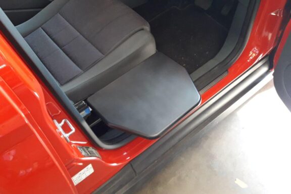 A Manual Transfer Plate is slotted into place next to and level with the driver's seat. A smooth black surface for sliding across sits at a height with the adjacent seat.