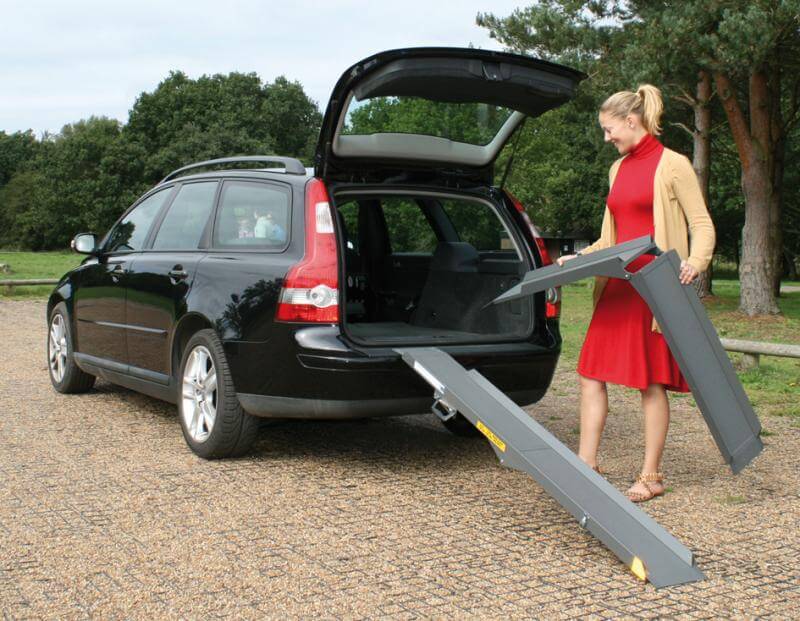 A woman is unfolding and attaching a pair of portable ramps to the open boot of an estate car.