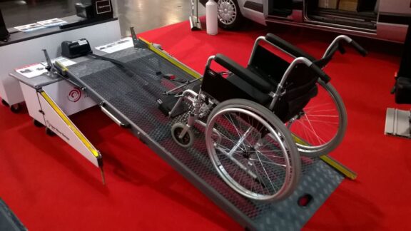 A Wheelchair Winch Assist is shown demonstrating how it lifts a wheelchair up a ramp. It is on a red carpeted area at a convention.