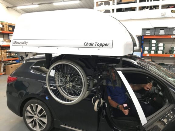 A folding wheelchair is lifted up to the white chair topper car roofbox.