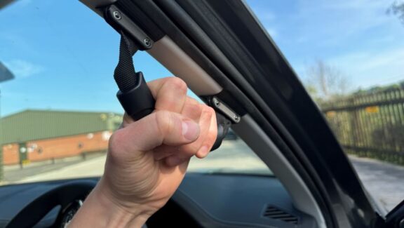 A black rubber handle is shown fitted to a car's A-pillar, with a hand grasping it.