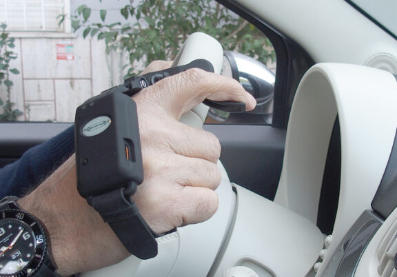 A black device, the Guidosimplex wireless trigger throttle, is strapped to a hand that is holding a cream car steering wheel. The device has a trigger being held by the hand which controls the car's acceleration wirelessly.