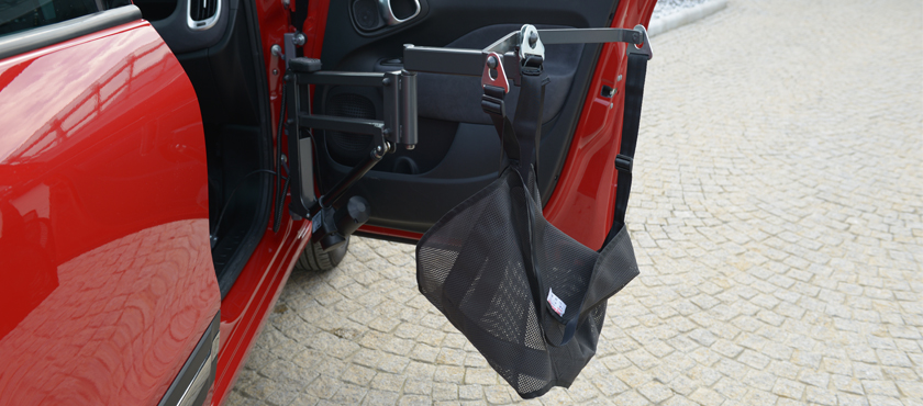 The Kivi person lift is shown empty, out of the passenger side door. It has a black mesh sling with three attachment points to a metal arm extended from the car.