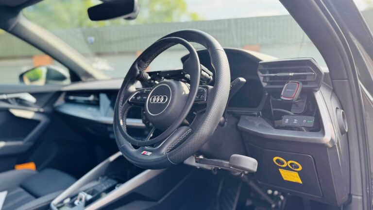 An Audi steering wheel with an over ring fitted to the front of it. A hand brake control is also fitted beneath the wheel on the right side.