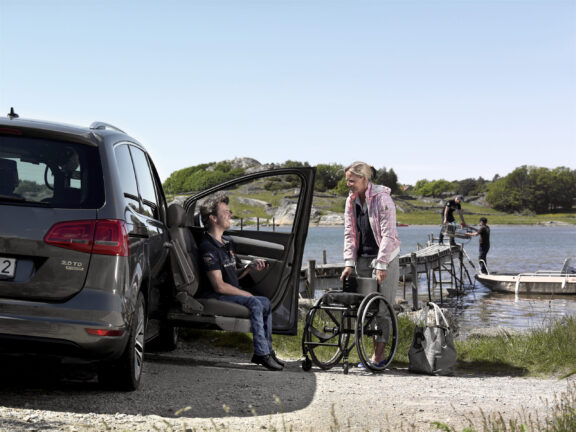 The Turny Evo swivel seat is extended out of a car by a wheelchair user preparing to transfer into his wheelchair against a sunny lakeside background.