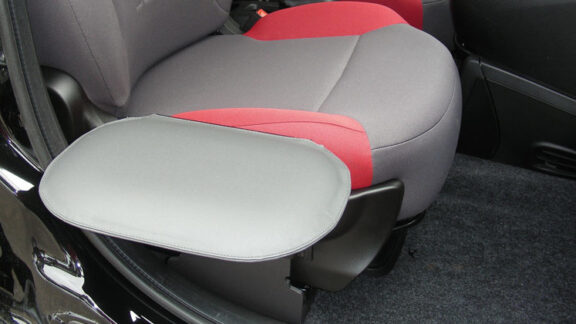 A SEG transfer plate is slotted into position next to a car seat. The plate is grey, matching the chair's fabric, and has some padding for comfort.