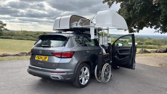 A rooftop box is open to extend a wheelchair winch down to the ground next to the open driver's side door. A folded wheelchair is attached.
