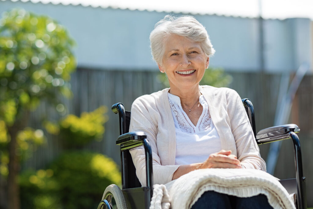 A woman is outside on a sunny day sitting in a wheelchair, smiling, with a blanket on her lap.